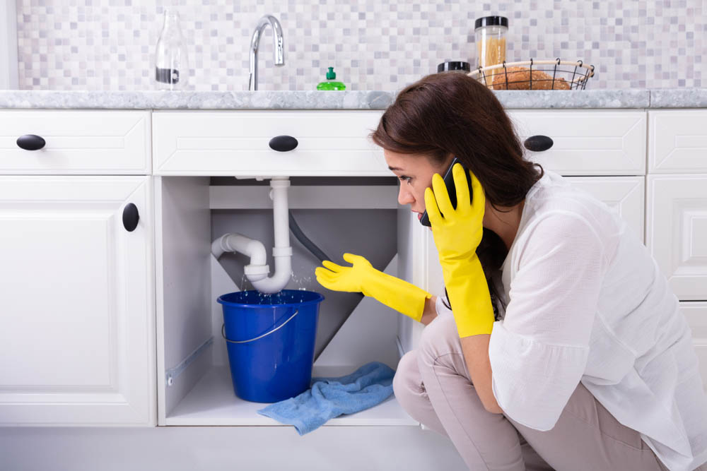 Woman Calling Plumber In Front Of Water Leaking From Sink Pipe